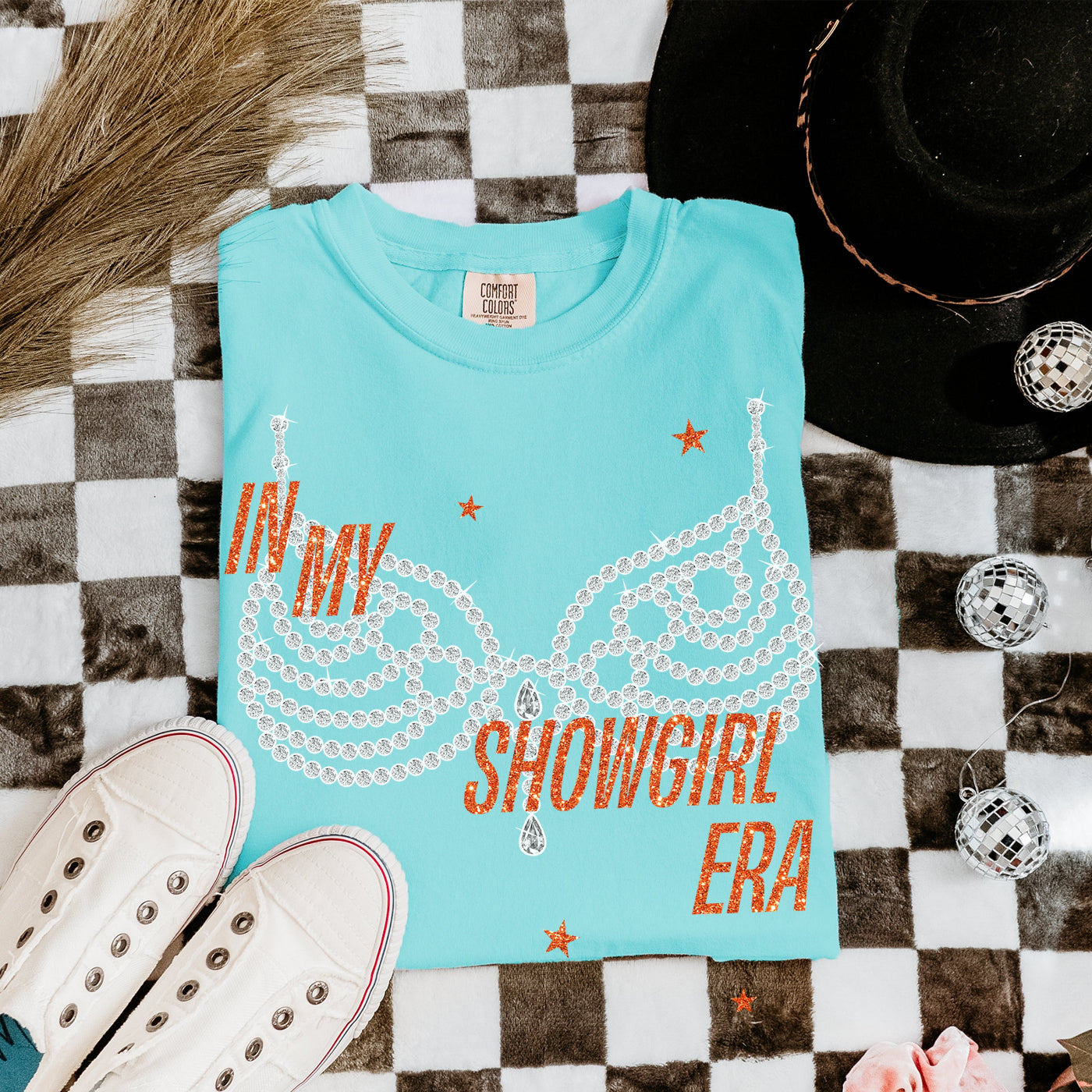 Turquoise t-shirt with 'IN MY SHOWGIRL ERA' text on a checkered surface with shoes and hat.